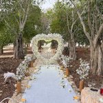 שער לב מפרחים לבנים עם שלט Marry Me בחורשת עצי זית, שביל לבן ונרות משני צדדיו