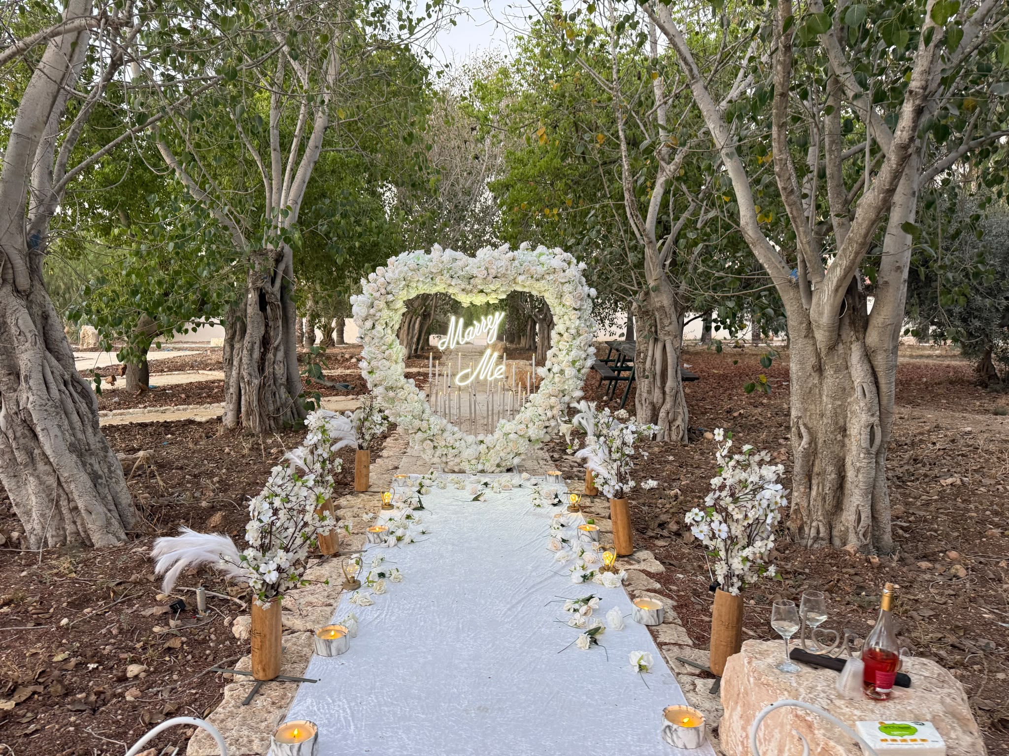 שער לב מפרחים לבנים עם שלט Marry Me בחורשת עצי זית, שביל לבן ונרות משני צדדיו