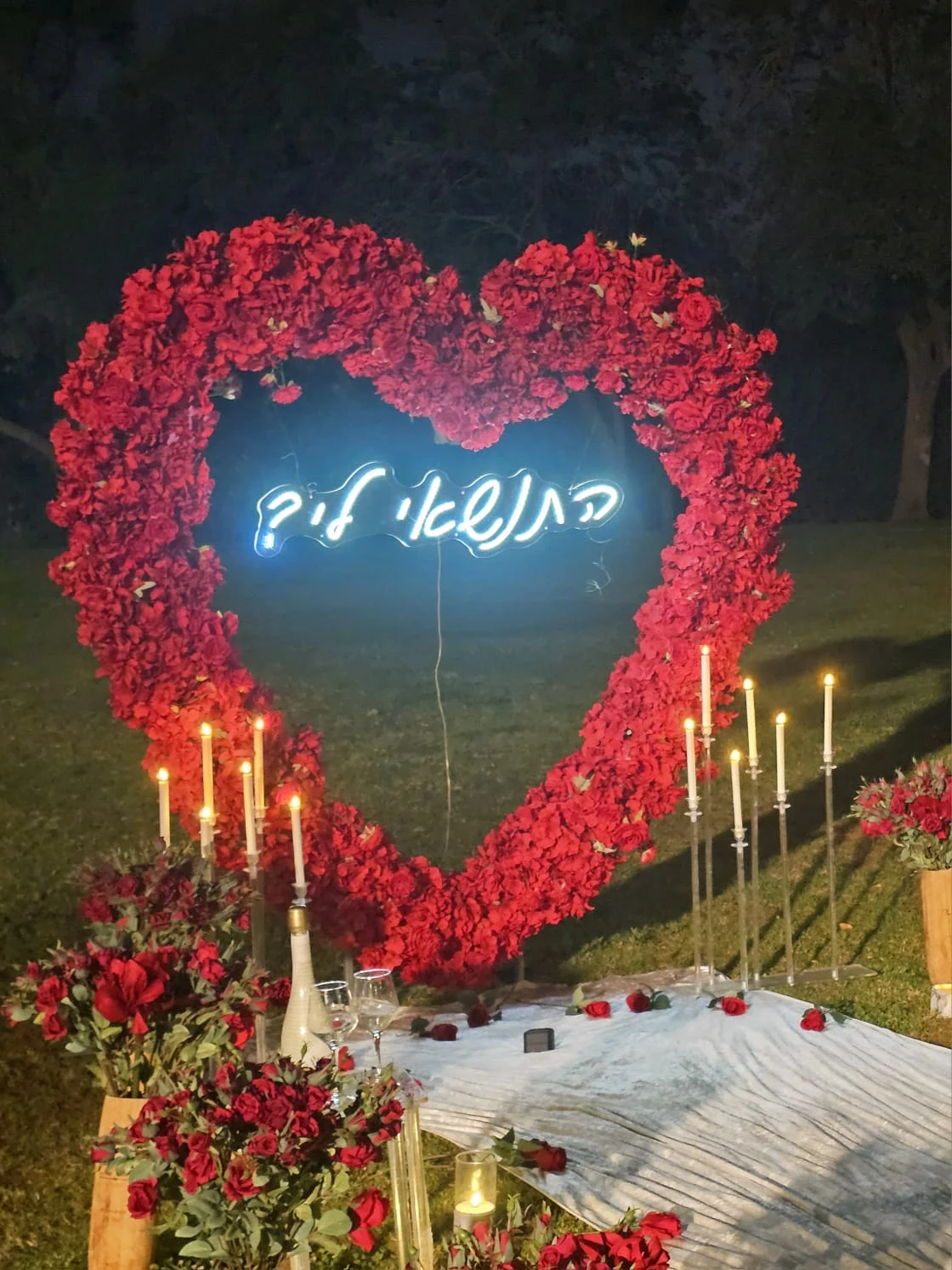 לב גדול מוורדים אדומים עם שלט ניאון "התנשאי לי?" מואר בלילה, נרות גבוהים, זרי ורדים אדומים וכוסות שמפניה